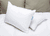 Two white Keeco Choice Hotels® Soft and Firm Polyester Pillows (Combo Pack, Includes 2)—one upright with a blue "CHOICE" tag, one flat—rest on a white bedspread with beige cushions behind, creating a cozy bedroom setting.