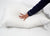 A hand with red nails in a navy blue pajama sleeve presses down on the Down Etc. 25% White Goose Down / 75% White Goose Feather Pillow, which rests on a white bed sheet with its side tag visible.