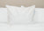 The Pillowtex Kyoto Pillow—half buckwheat, half polyester—is displayed upright on a white quilted comforter, its tag visible, against a soft beige wall for a clean, minimal look.