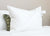 The Manchester Mills Down Dreams Classic Firm Pillow (formerly Classic Too) with a visible tag rests on a white bed. Behind it, a smaller green pillow with a black floral pattern is partially visible against a plain beige wall.