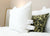 A close-up shows a neatly made bed featuring the Hollander Eco-Smart Down Alternative Pillow | Made in USA with tags, in front of an olive green pillow with black floral designs, both on crisp white bedding and a soft light in the background.