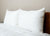 Two large white pillows in Pillowtex Bamboo Pillow Covers by Pillowtex rest against a light beige wall on a wooden headboard atop a neatly made bed with a white quilted comforter, creating a clean and inviting look.
