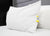 A large white Martex Brentwood Gold Pillow, featured at many hotels, rests against another pillow on a neatly made bed with white sheets and a gray upholstered headboard, creating a hotel-style look in an uncluttered setting.
