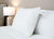 A close-up shows a neatly made bed with a white Keeco Best Western® Aurora Pillow | Medium Polyester Pillow, its care label visible and propped against the headboard. A white table lamp and mirror enhance the clean, bright setting.