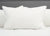 The Down Etc. Rhapsody Wrap Down/Feather Pillow, featured at many Hyatt® Hotels, sits upright with its soft, inviting surface and a small fabric tag on a neatly made white bed against a light grey upholstered headboard.