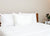 A neatly made bed features three Pacific Coast Feather Tria Down & Feather Pillows, as seen at Ritz-Carlton® Hotels, with a wooden headboard. A nightstand holds a plant in a white pot and an alarm clock against a light beige wall.