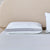 A neatly made bed features the Carpenter Indulgence® Synthetic Down Pillow for side sleepers in a striped case with "Indulgence" on its edge, set against a beige upholstered headboard and finished with a plain white bedsheet.