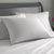 A neatly made bed features Hollander's Holiday Inn® Soft and Firm Polyester Pillow Combo Pack—two large, supportive pillows with dark piping—against a gray headboard. A magenta flower in a vase adds a touch of style to the scene.